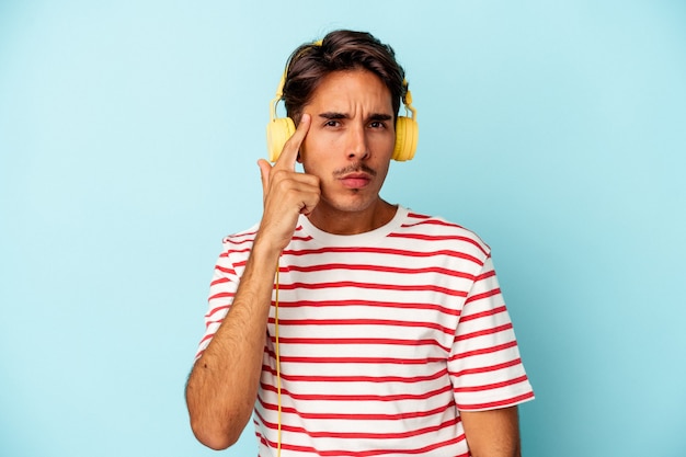 Giovane uomo di razza mista che ascolta musica isolato su sfondo blu che punta il tempio con il dito, pensando, concentrato su un compito.