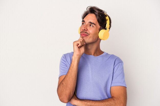 Giovane uomo di razza mista che ascolta musica isolato su sfondo blu guardando di traverso con espressione dubbiosa e scettica.