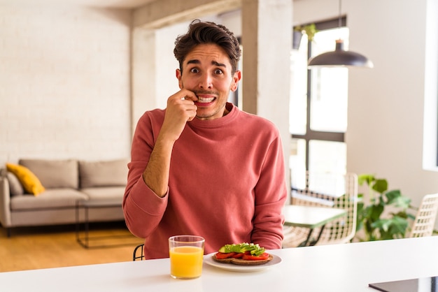 Giovane uomo di razza mista che fa colazione in cucina mangiandosi le unghie, nervoso e molto ansioso.