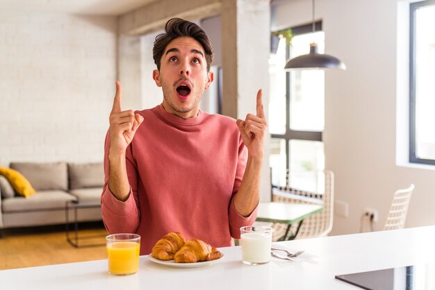 Giovane uomo di razza mista che fa colazione in una cucina la mattina che punta al rialzo con la bocca aperta.