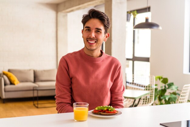 Giovane uomo di razza mista che fa colazione nella sua cucina felice, sorridente e allegro.