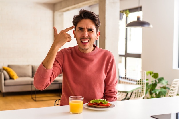 Giovane uomo di razza mista che fa colazione nella sua cucina mostrando un gesto di delusione con l'indice.