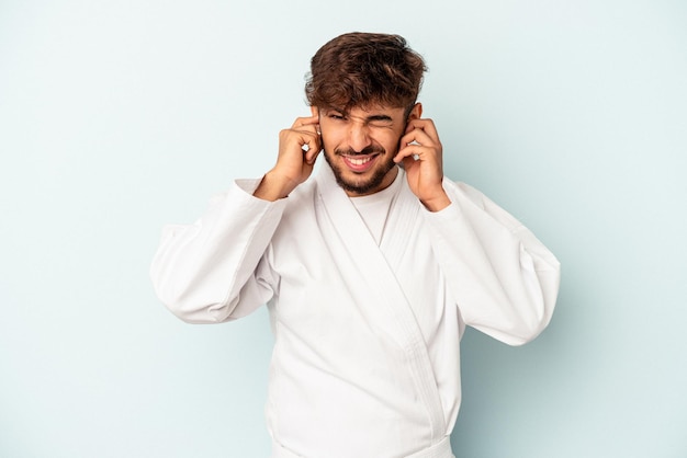 Giovane uomo di razza mista che fa karate isolato su sfondo blu che copre le orecchie con le mani.