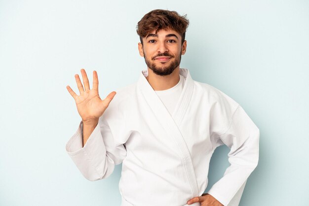 Giovane uomo di razza mista che fa karate isolato su sfondo blu sorridente allegro che mostra il numero cinque con le dita.