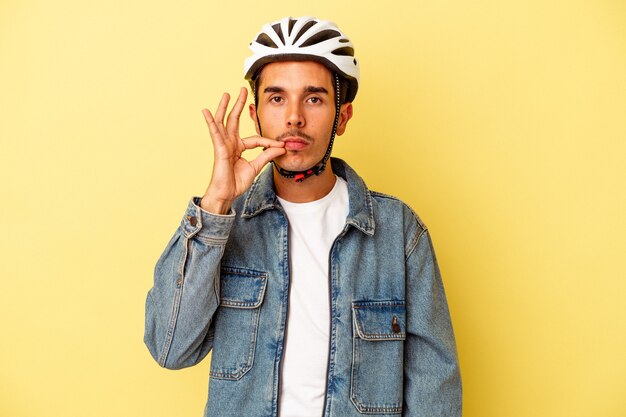 Giovane uomo di razza mista che indossa una bici con casco isolata su sfondo giallo con le dita sulle labbra mantenendo un segreto.