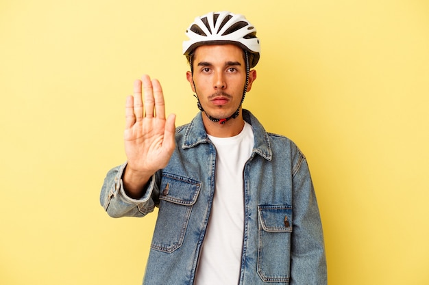 Giovane uomo di razza mista che indossa una bici con casco isolata su sfondo giallo in piedi con la mano tesa che mostra il segnale di stop, impedendoti.