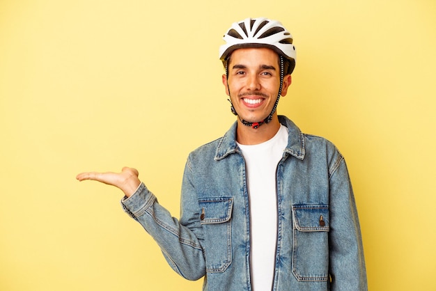 Giovane uomo di razza mista che indossa una bici da casco isolata su sfondo giallo che mostra uno spazio di copia su un palmo e tiene un'altra mano sulla vita.