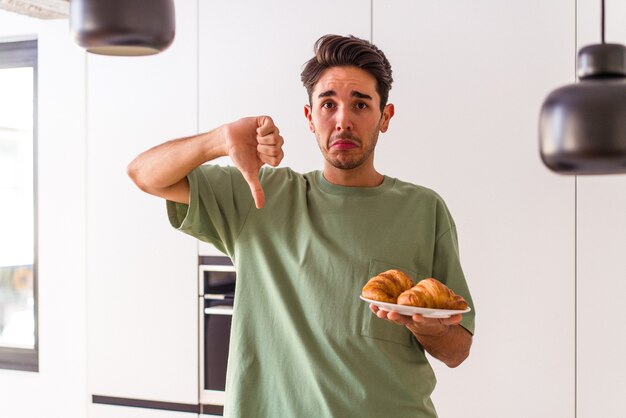 Giovane uomo di razza mista che mangia croissant in una cucina la mattina mostrando un gesto di antipatia, pollice verso. Concetto di disaccordo.