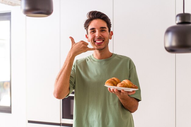 Giovane uomo di razza mista che mangia croissant in una cucina la mattina mostrando un gesto di telefonata con le dita.