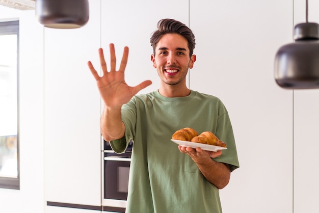 Giovane uomo di razza mista che mangia croissant in una cucina la mattina sorridente allegro che mostra il numero cinque con le dita.