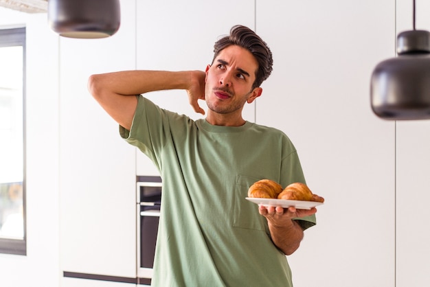 Giovane uomo di razza mista che mangia croissant in una cucina la mattina toccando la nuca, pensando e facendo una scelta.