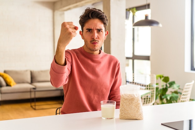 Giovane uomo di razza mista che mangia farina d'avena e latte per colazione nella sua cucina che mostra il pugno alla telecamera, espressione facciale aggressiva.