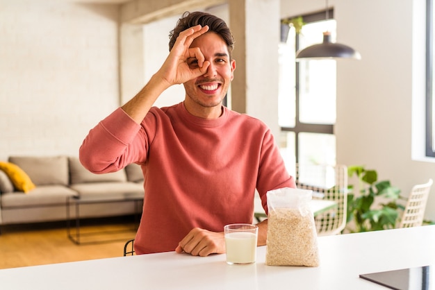 Giovane uomo di razza mista che mangia farina d'avena e latte per colazione nella sua cucina eccitato mantenendo il gesto ok sull'occhio.