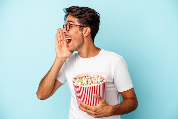 Giovane uomo di razza mista che mangia popcorn isolati su sfondo blu gridando e tenendo il palmo vicino alla bocca aperta.