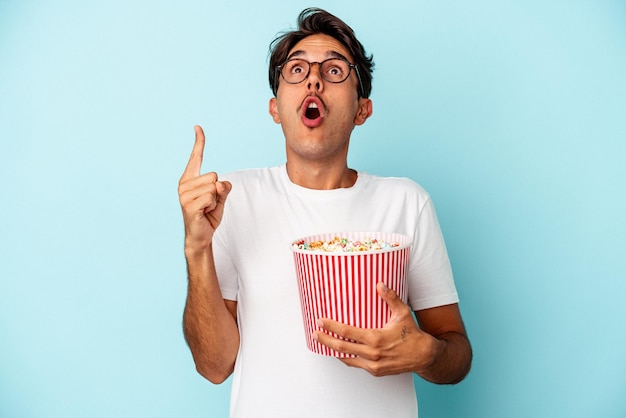 Giovane uomo di razza mista che mangia popcorn isolato su sfondo blu rivolto verso l'alto con la bocca aperta.