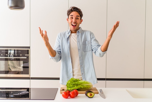 Giovane uomo di razza mista che prepara un'insalata per pranzo ricevendo una piacevole sorpresa, eccitato e alzando le mani.