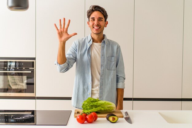 Giovane uomo di razza mista che prepara un'insalata per pranzo sorridente allegro che mostra il numero cinque con le dita.