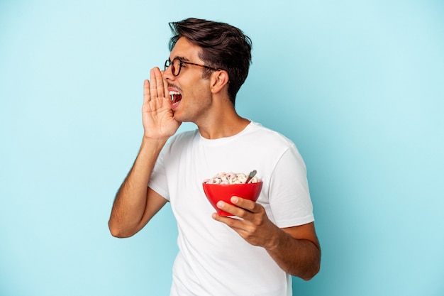 Giovane uomo di razza mista che tiene i cereali isolati su sfondo blu gridando e tenendo il palmo vicino alla bocca aperta.