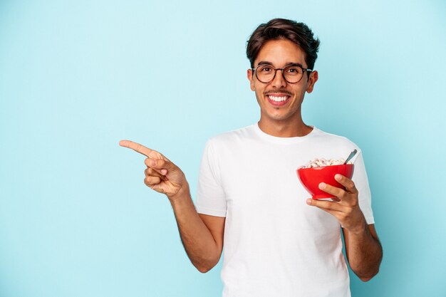 Giovane uomo di razza mista che tiene i cereali isolati su sfondo blu sorridendo e indicando da parte, mostrando qualcosa in uno spazio vuoto.