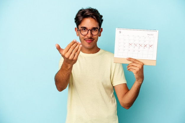 Giovane uomo di razza mista che tiene il calendario isolato su sfondo blu che punta con il dito su di te come se invitasse ad avvicinarsi.