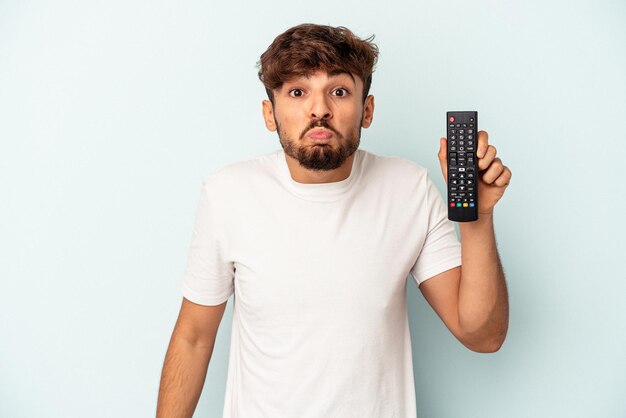 Giovane uomo di razza mista che tiene in mano un controller tv isolato su sfondo blu alza le spalle e apre gli occhi confusi.