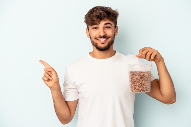 Giovane uomo di razza mista che tiene un cibo per uccelli isolato su sfondo blu sorridente e puntato da parte, mostrando qualcosa in uno spazio vuoto.