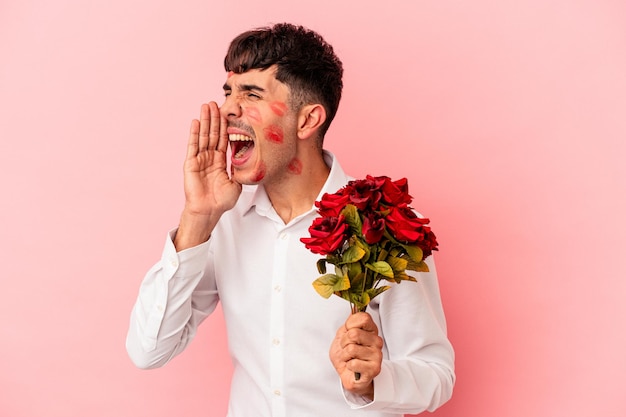 Giovane uomo di razza mista che tiene un mazzo di fiori isolato su sfondo rosa che grida e tiene il palmo vicino alla bocca aperta.