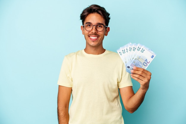 Giovane uomo di razza mista con banconote isolate su sfondo blu felice, sorridente e allegro.