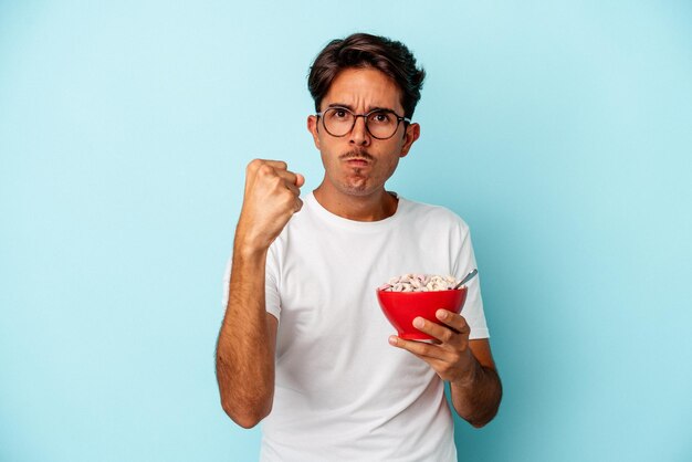 Giovane uomo di razza mista con cereali isolati su sfondo blu che mostra il pugno alla telecamera, espressione facciale aggressiva.