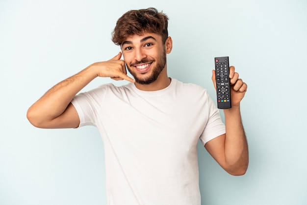 Giovane uomo di razza mista in possesso di un controller tv isolato su sfondo blu che mostra un gesto di chiamata di telefonia mobile con le dita.