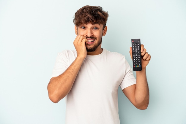 Giovane uomo di razza mista in possesso di un controller tv isolato su sfondo blu che si morde le unghie, nervoso e molto ansioso.