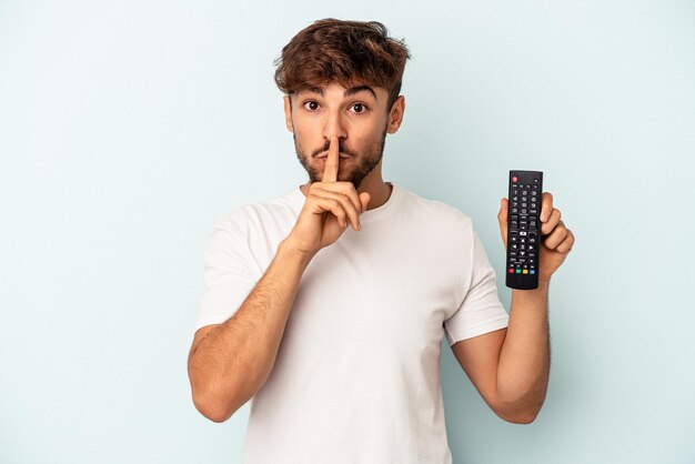 Giovane uomo di razza mista in possesso di un controller tv isolato su sfondo blu mantenendo un segreto o chiedendo silenzio.