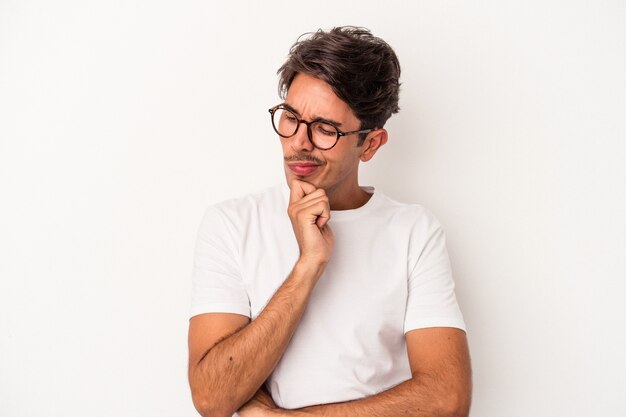 Giovane uomo di razza mista isolato su sfondo bianco guardando lateralmente con espressione dubbiosa e scettica.