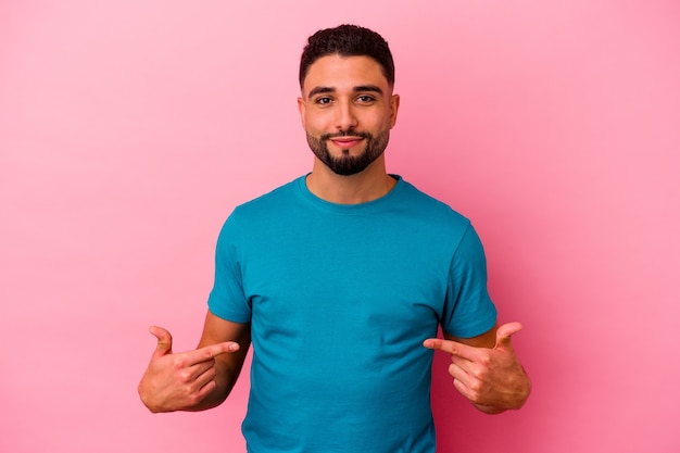 Giovane uomo di razza mista isolato su sfondo rosa persona che punta a mano uno spazio di copia camicia, orgoglioso e fiducioso