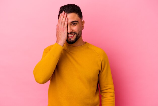 Giovane uomo di razza mista isolato sul muro rosa divertendosi a coprire metà del viso con il palmo.