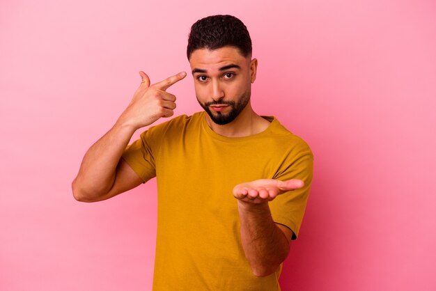 Giovane uomo di razza mista isolato sulla parete rosa che tiene e che mostra un prodotto a portata di mano.