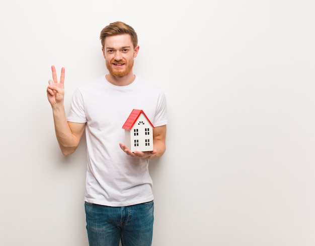 Giovane uomo di redhead che mostra numero due. In possesso di un modello di casa.