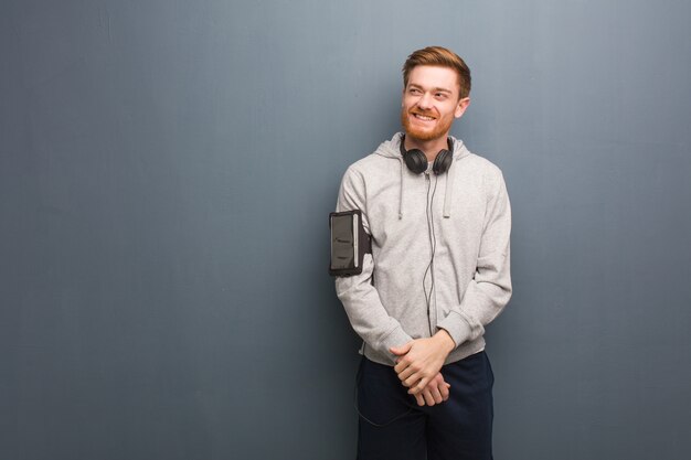 Giovane uomo di redhead di forma fisica che sorride sicuro e che attraversa armi, guardanti su