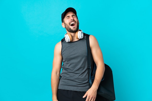 Giovane uomo di sport con borsa sportiva isolata sulla parete blu che ride