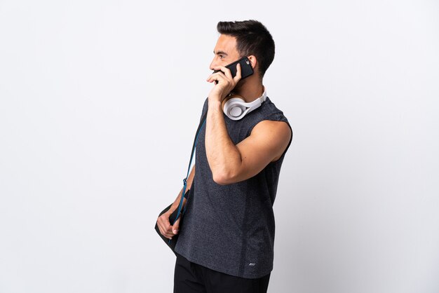 Giovane uomo di sport con borsa sportiva isolato su sfondo bianco mantenendo una conversazione con il telefono cellulare con qualcuno