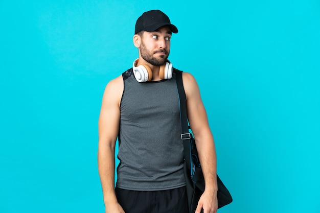 Giovane uomo di sport con borsa sportiva isolato su sfondo blu facendo dubbi gesto guardando lato