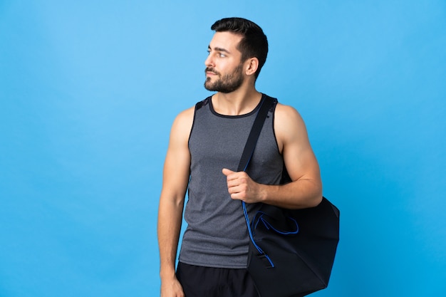 Giovane uomo di sport con la borsa di sport isolata sulla parete blu che guarda al lato