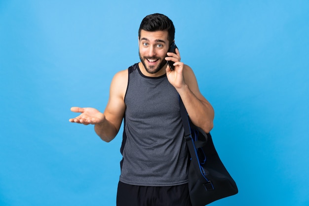 Giovane uomo di sport con la borsa di sport isolata sulla parete blu che mantiene una conversazione con il telefono cellulare con qualcuno