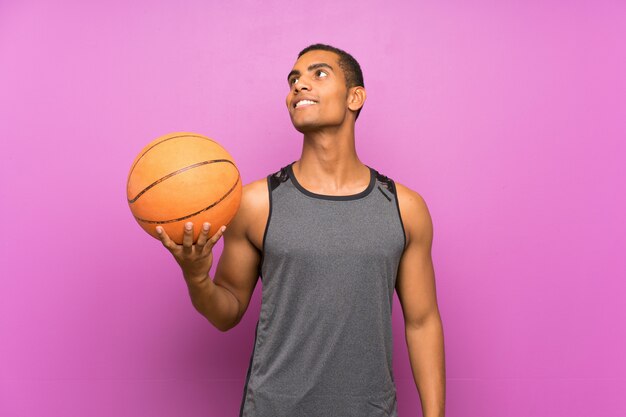 Giovane uomo di sport con la palla di pallacanestro sopra la parete porpora isolata che cerca mentre sorridendo