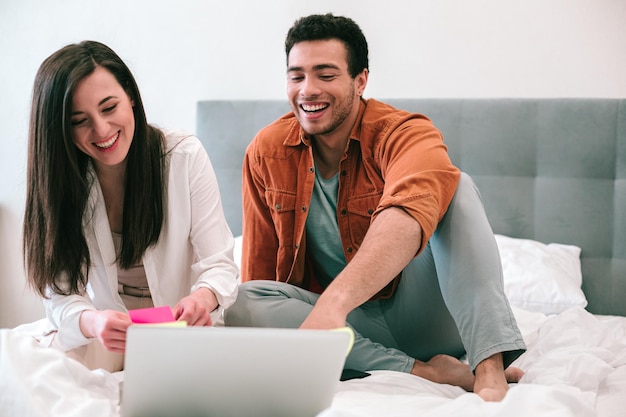 Giovane uomo e donna contenti seduti su un letto comodo con un laptop davanti a loro e sorridenti mentre lo guardano