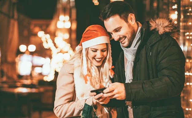 Giovane uomo e donna in piedi per strada alla vigilia di Natale e divertirsi guardando lo smartphone.