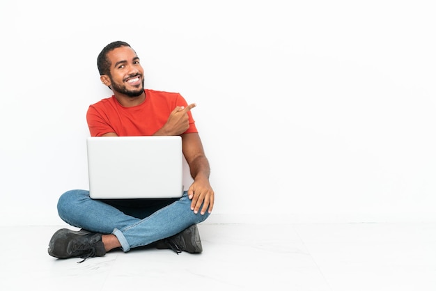 Giovane uomo ecuadoriano con un computer portatile seduto sul pavimento isolato su sfondo bianco che punta indietro