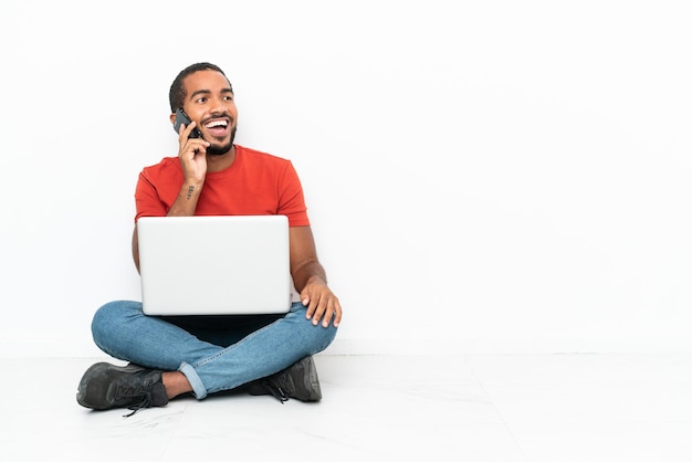 Giovane uomo ecuadoriano con un computer portatile seduto sul pavimento isolato su sfondo bianco mantenendo una conversazione con il telefono cellulare