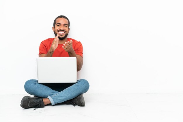 Giovane uomo ecuadoriano con un laptop seduto sul pavimento isolato su sfondo bianco che applaude dopo la presentazione in una conferenza