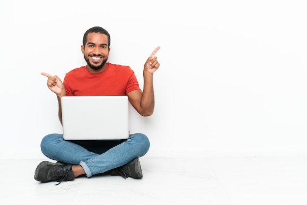 Giovane uomo ecuadoriano con un laptop seduto sul pavimento isolato su sfondo bianco che punta il dito verso i laterali e felice